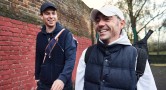 Two men smile while walking side by side beside a wall at a tennis club.